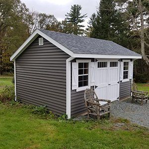Gray-sided shed with white trim and doors, topped by a dark roof, set in a grassy yard. Two wooden chairs sit nearby.