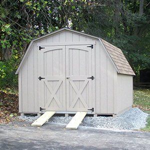 Tan barn-style shed with double doors, black hinges, and two wooden ramps on a gravel base.