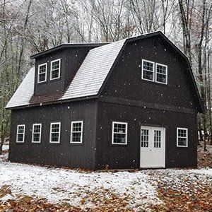 Dark barn with white-trimmed windows and doors, snow-covered roof, snowy setting.