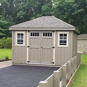 Tan shed with gray roof, white trim, and double doors, set on a paved area, and small fence.