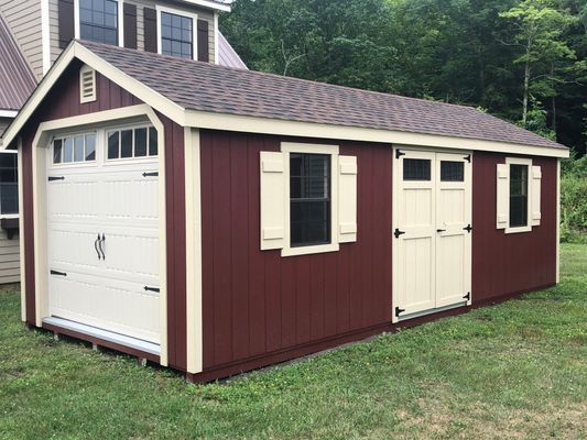 Red shed with garage door and side door, beige trim, windows, and brown roof on green grass.