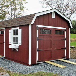Red barn-style shed with white trim, door ramp, and window with shutters set on gravel, in a yard.
