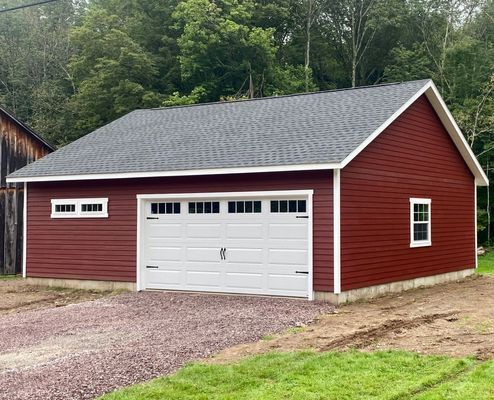 Red-sided garage with white door, windows, and a gray roof sits on a gravel driveway, surrounded by green grass and trees.