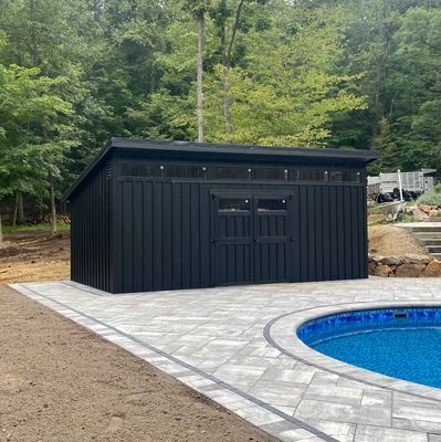 Black shed with double doors next to a pool. Dark siding, gray patio, and trees in the background.