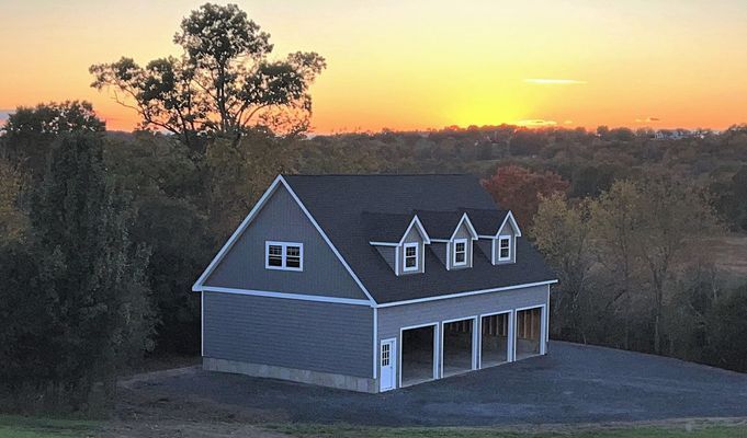 Three-bay garage with dormers, gray siding, set against a sunset over a wooded landscape.