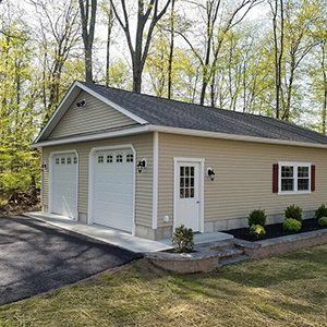 Tan two-car garage with white doors, entry door, and red window shutters, set in a wooded area.