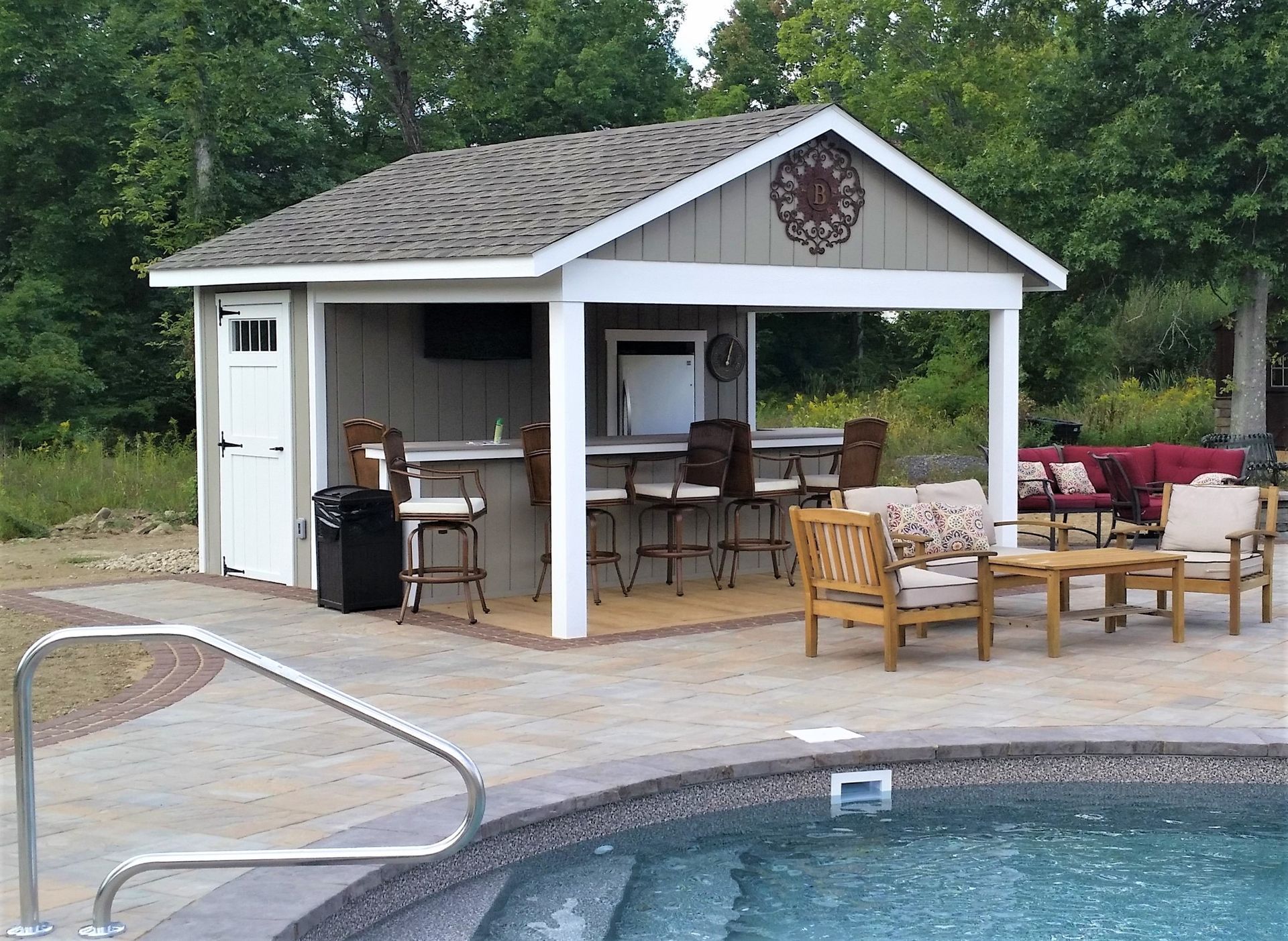 Outdoor bar with seating by a pool; tan and white structure, chairs, and a decorative wall piece.