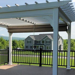 Pergola on deck overlooking a house and green landscape. White frame, brown awning.