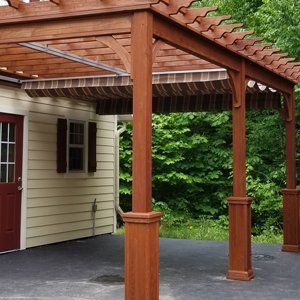 Wooden pergola with awning over a patio, next to a house with a red door.