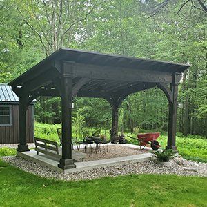 Dark stained wooden pergola with seating and red wheelbarrow on concrete pad in a yard.