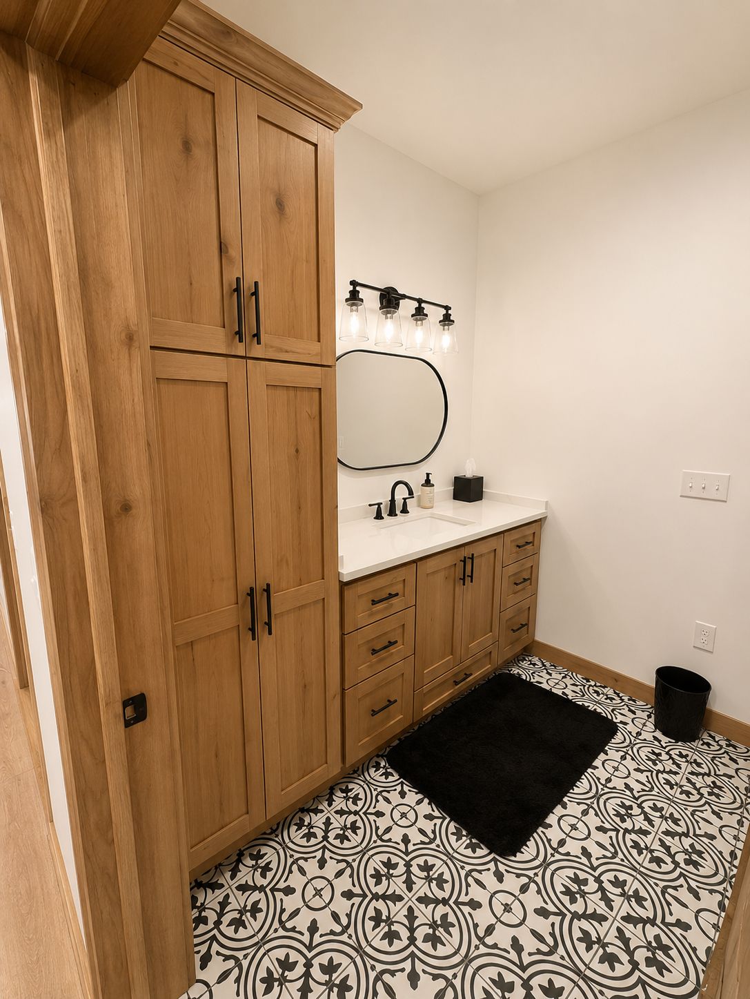 Bathroom with wooden cabinets, white vanity, oval mirror, black rug, and patterned tile floor