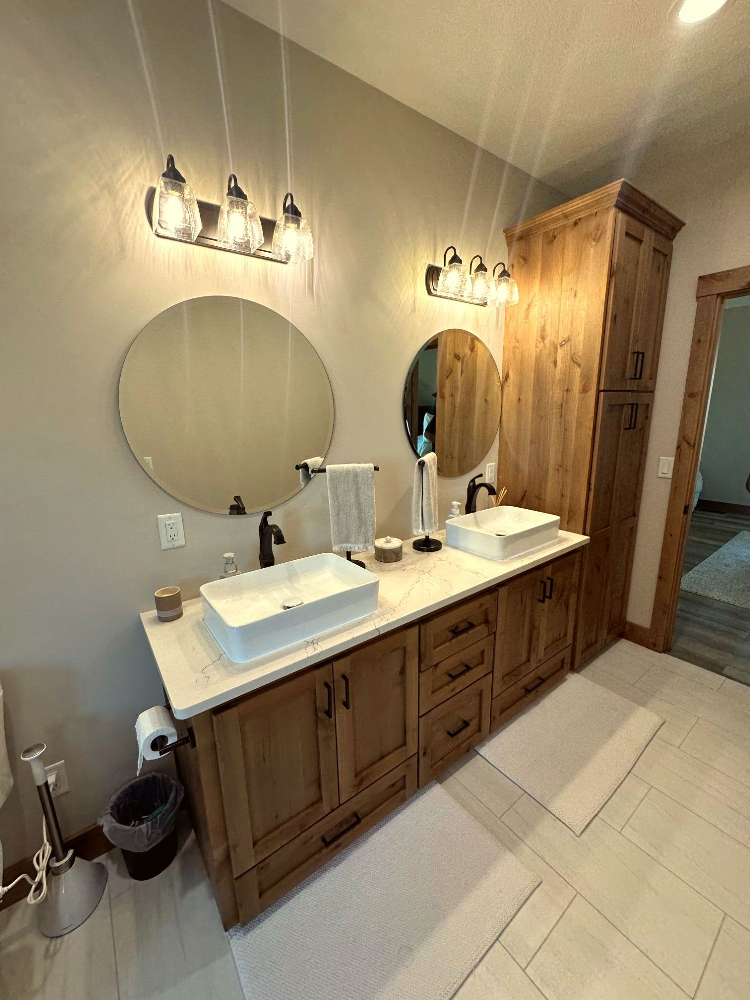 A double-sink bathroom vanity with rustic wood cabinets, white countertops, round mirrors, and glass pendant lights.