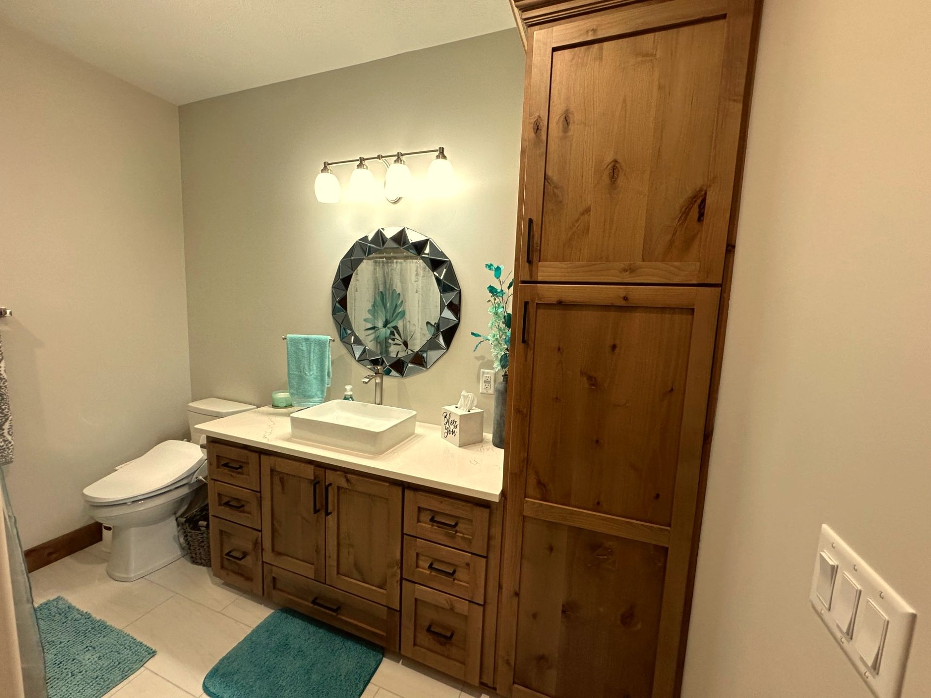 A rustic bathroom featuring a wooden vanity, a tall matching cabinet, a white vessel sink, and a circular mirror.
