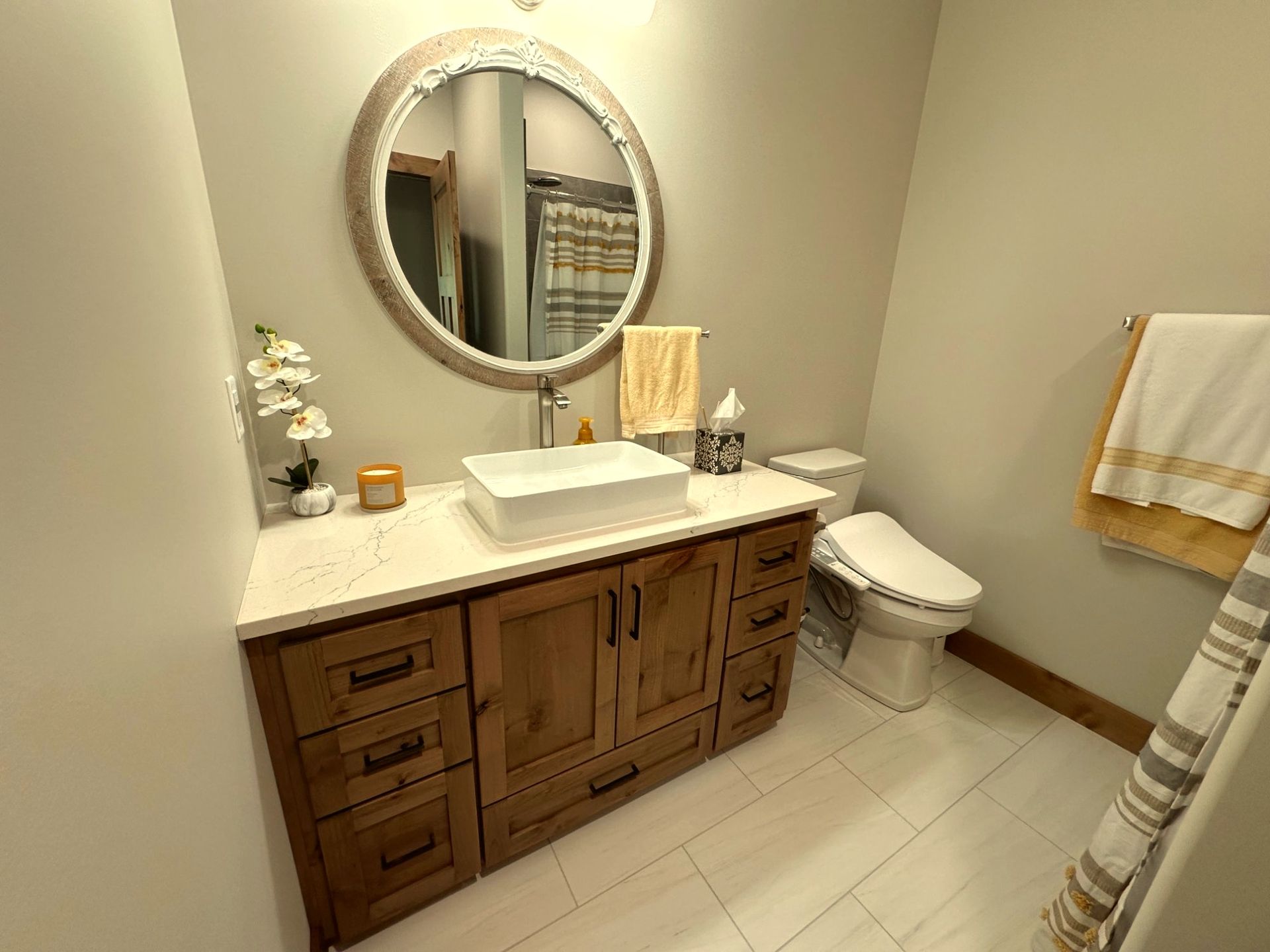 A modern bathroom featuring a wooden vanity with a white vessel sink, a circular mirror, and a toilet on white tile floor.