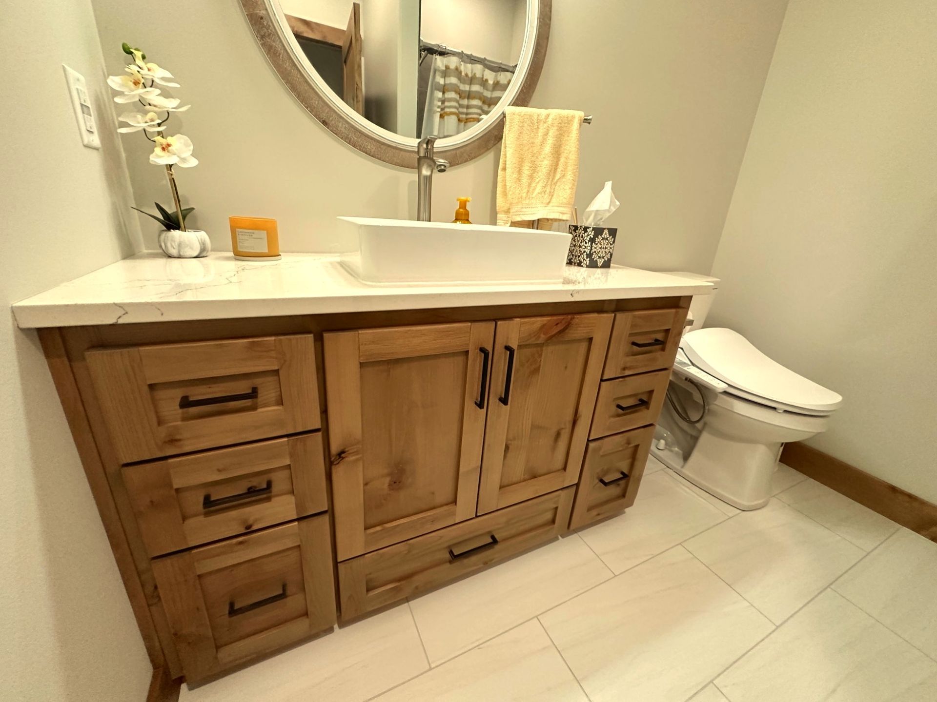 A rustic wooden bathroom vanity with a white rectangular sink, round mirror, and a toilet on tiled flooring.