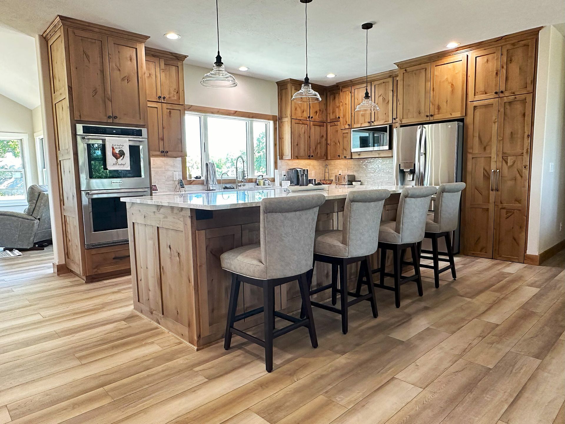 A rustic kitchen with wood cabinets, a marble-topped island, light gray bar stools, and warm wood flooring.