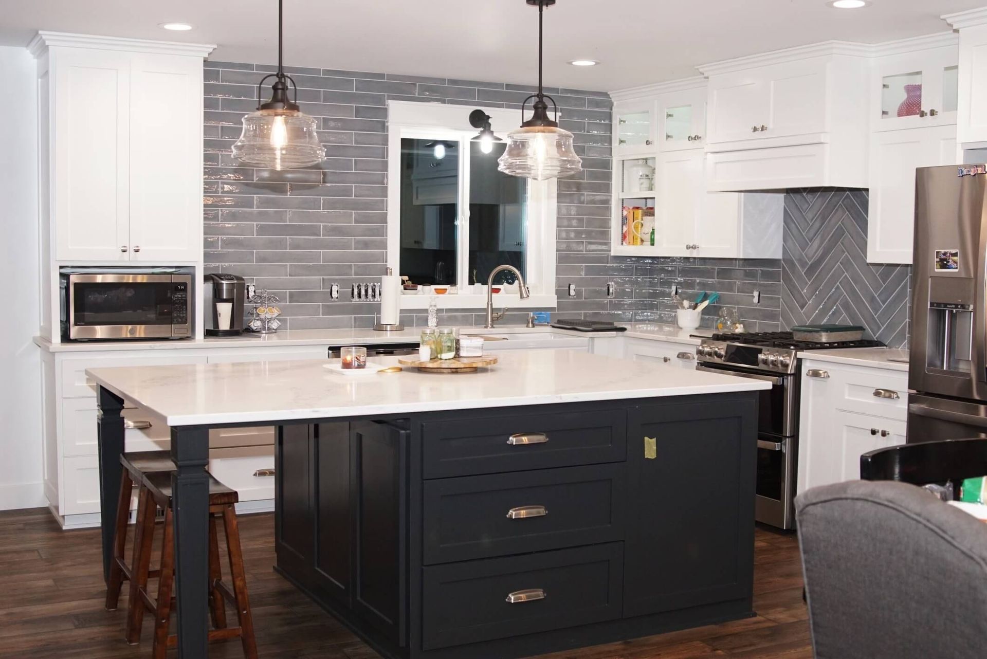 A kitchen featuring white cabinets, a dark blue island with bar stools, gray tiled walls, and two hanging pendant lights.