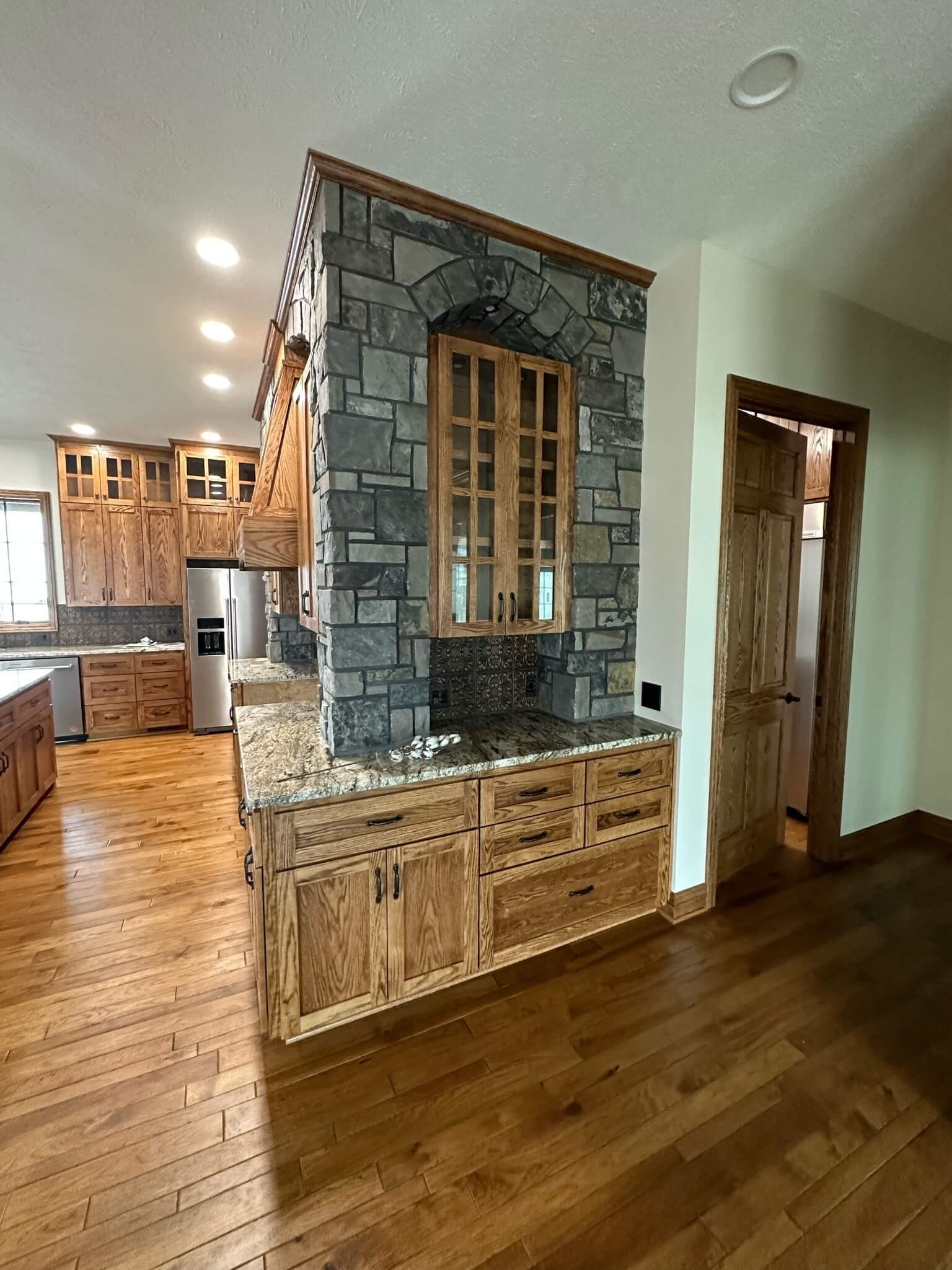 Rustic kitchen with a stone-accented cabinet tower, natural wood cabinetry, granite countertops, and hardwood floors.
