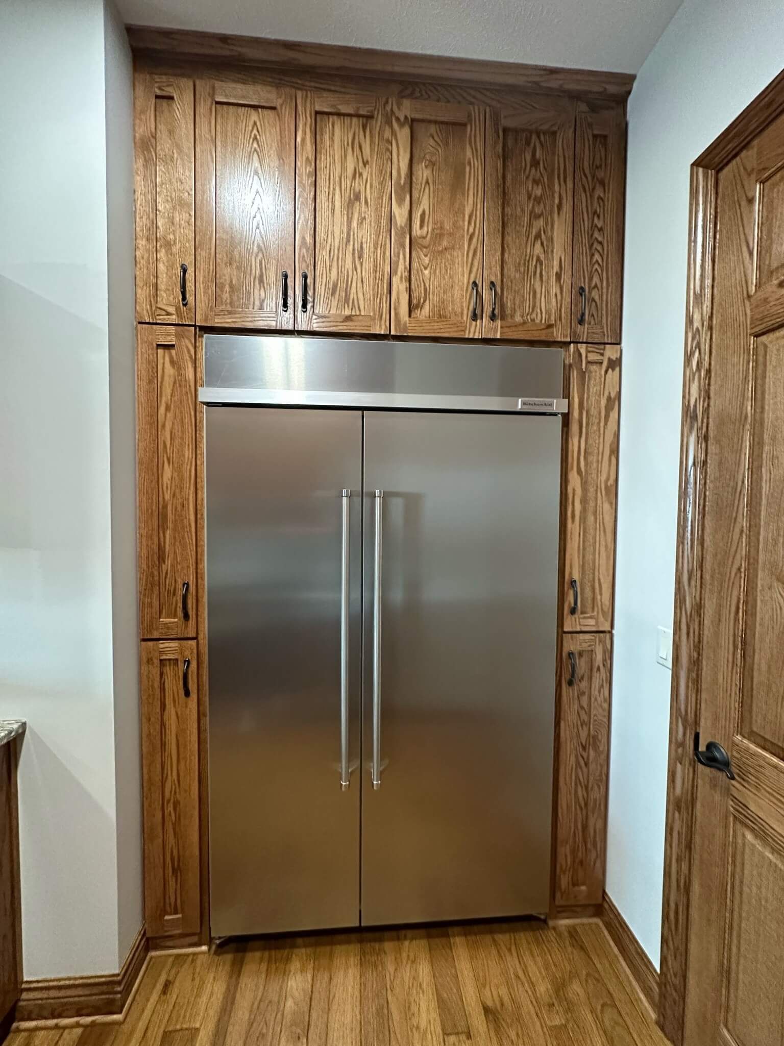 A stainless steel double-door refrigerator encased in matching wood-paneled cabinetry with storage above and to the sides.
