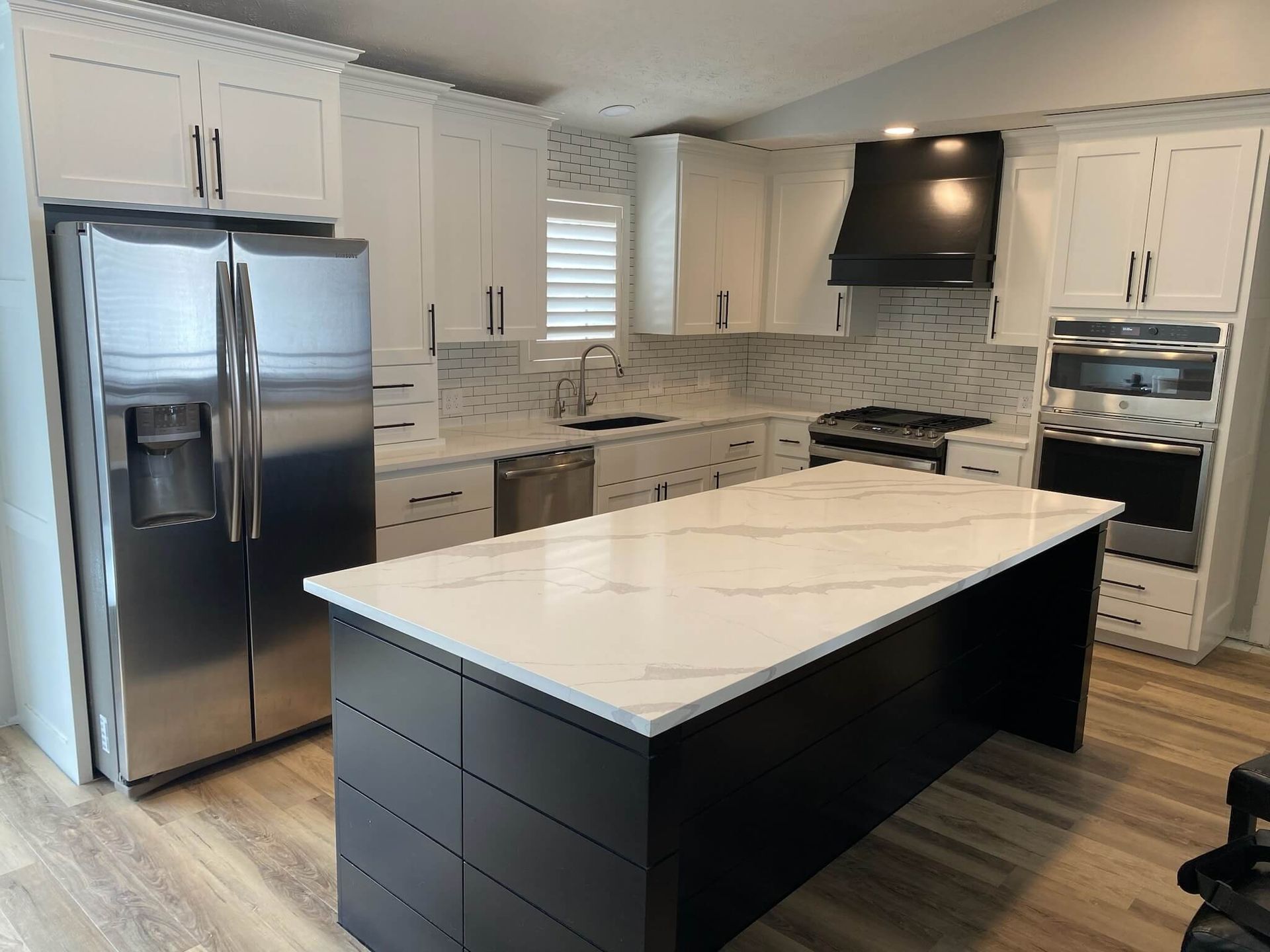 A bright kitchen with white cabinets, stainless steel appliances, a black range hood, and a dark island with marble top.