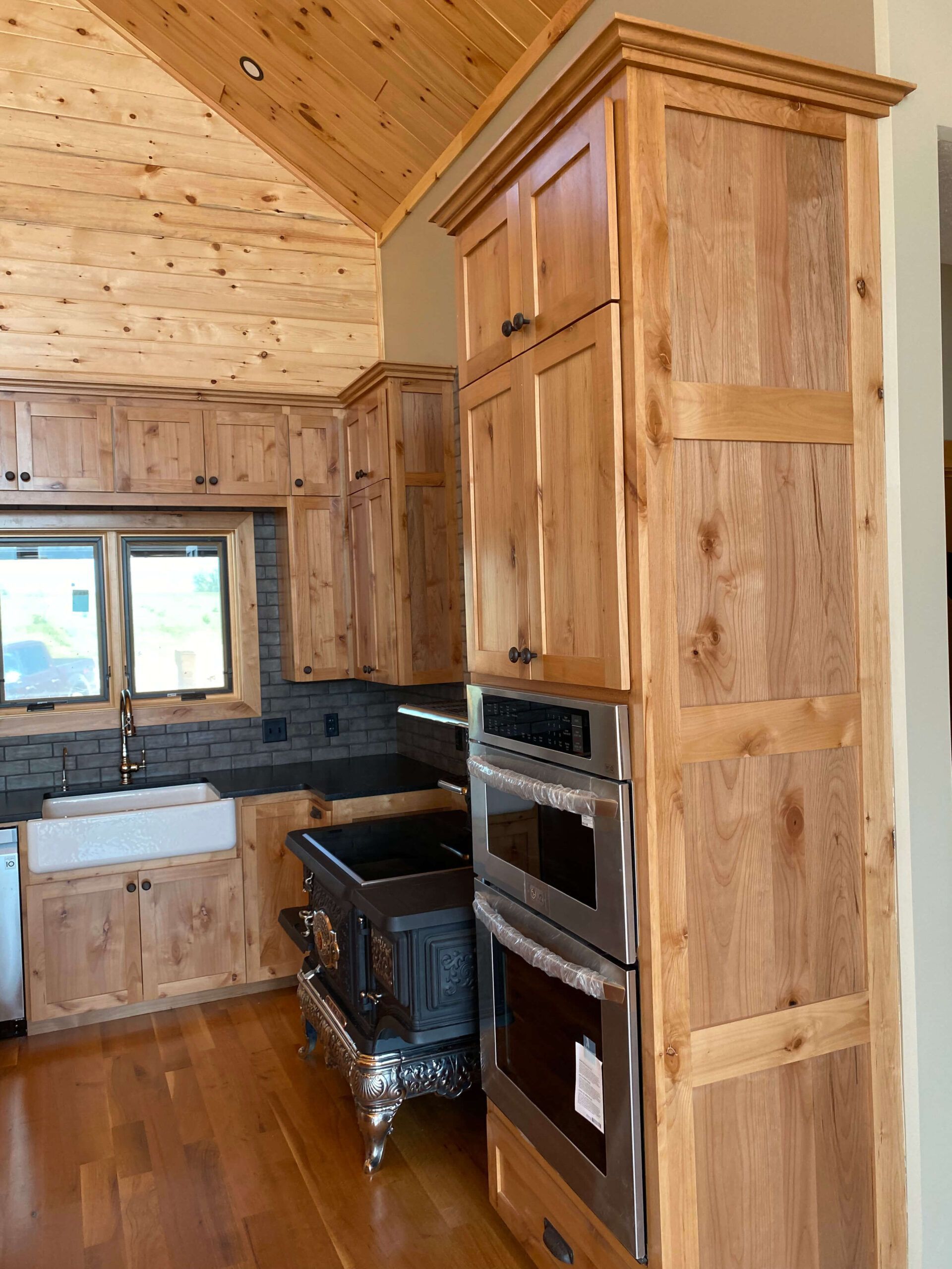 A rustic kitchen featuring natural wood cabinets, a stainless steel double oven, and a wood-burning stove under a high ceiling.