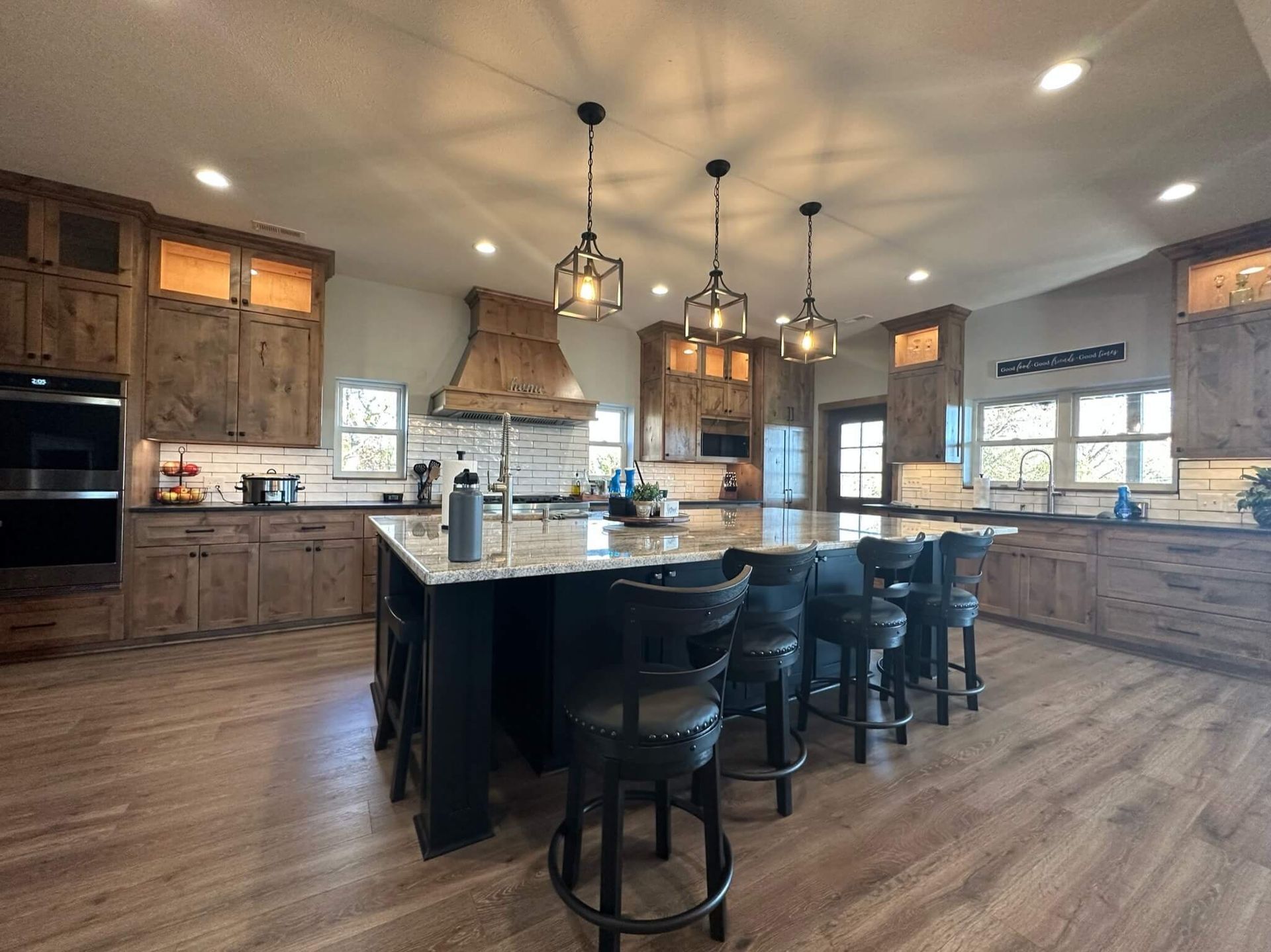 A modern kitchen with wooden cabinets, a large central island with black stools, and pendant lighting.