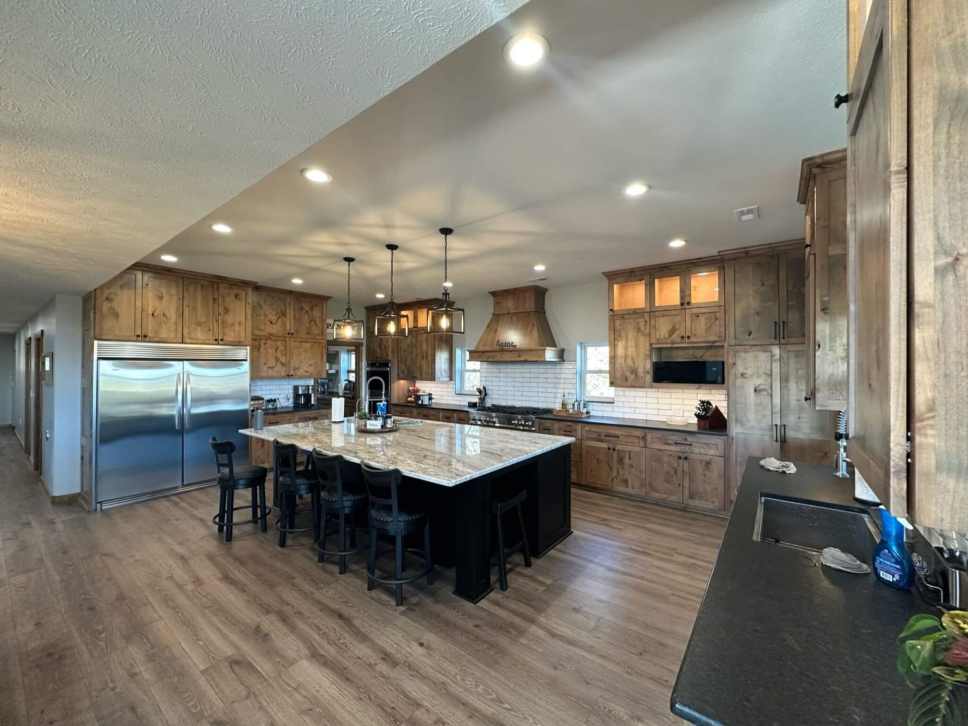 A modern kitchen features wood cabinets, a large granite-topped island with black stools, and stainless steel appliances.