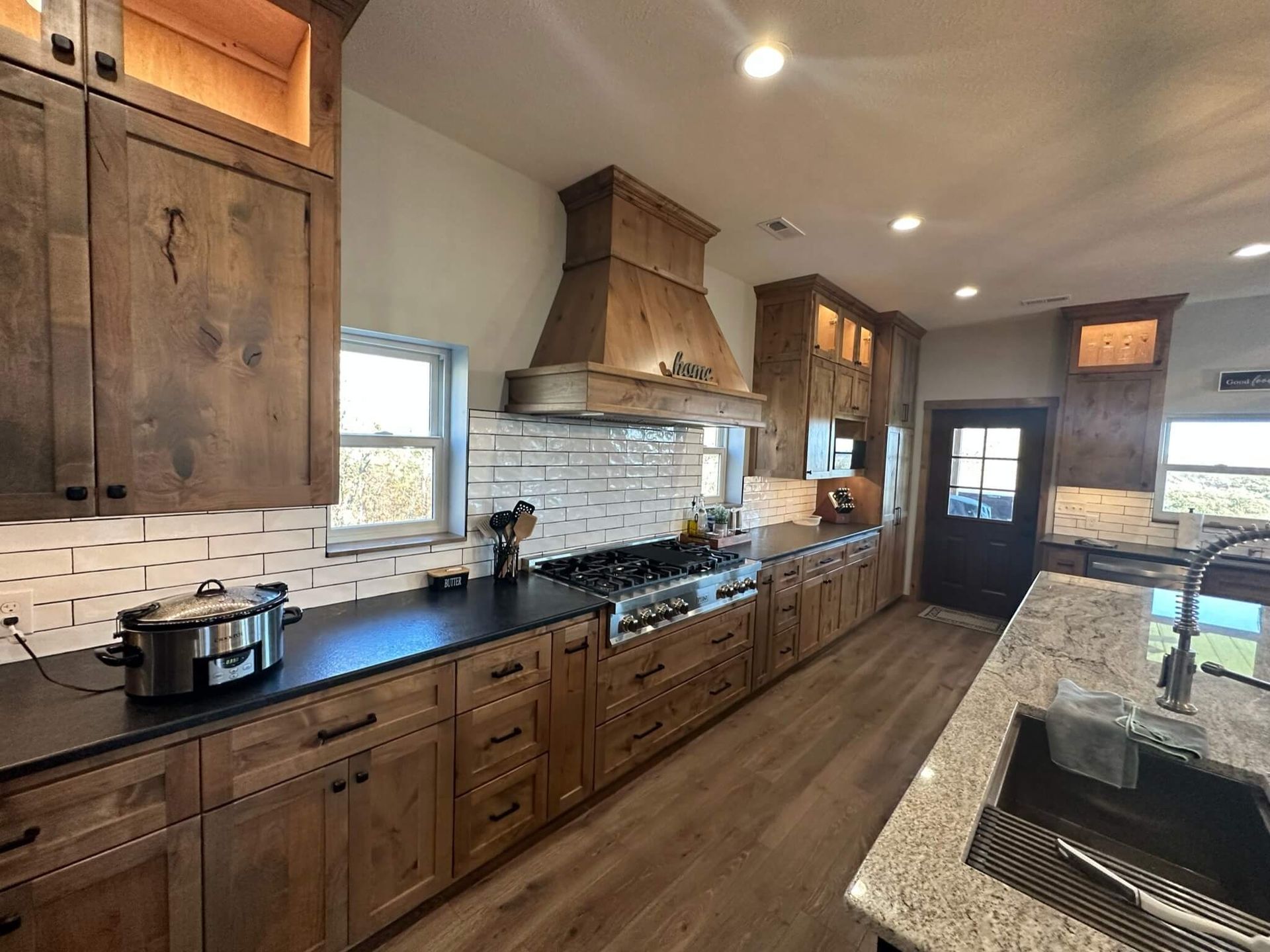 A spacious kitchen with rustic wooden cabinets, dark countertops, light flooring, a large range hood, and a kitchen island.