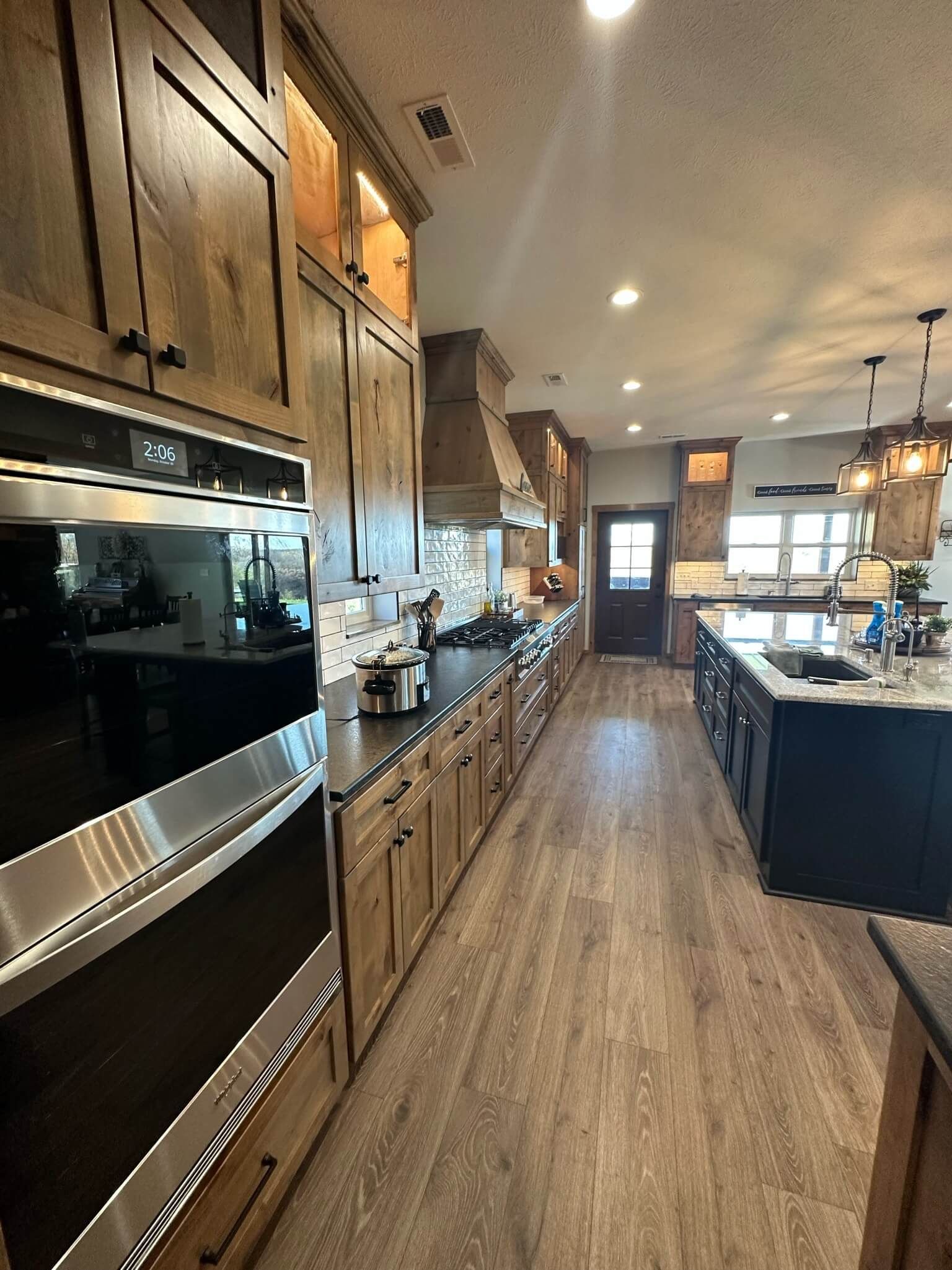 A long kitchen with wooden cabinets, dark appliances, a central island, and wood-patterned flooring.