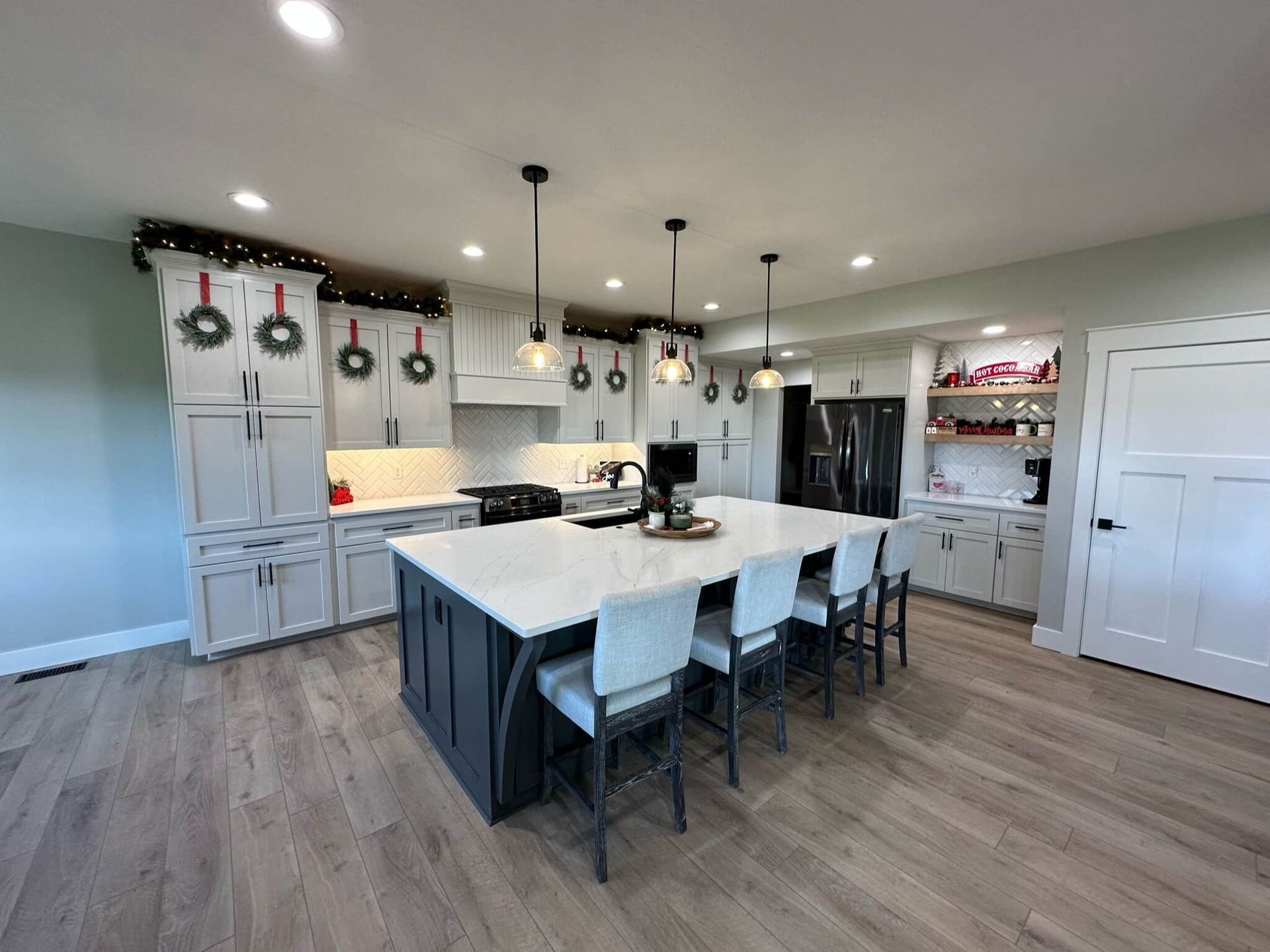 Modern kitchen with a dark grey island, white cabinets, light wood floors, and holiday wreaths on the cabinetry.