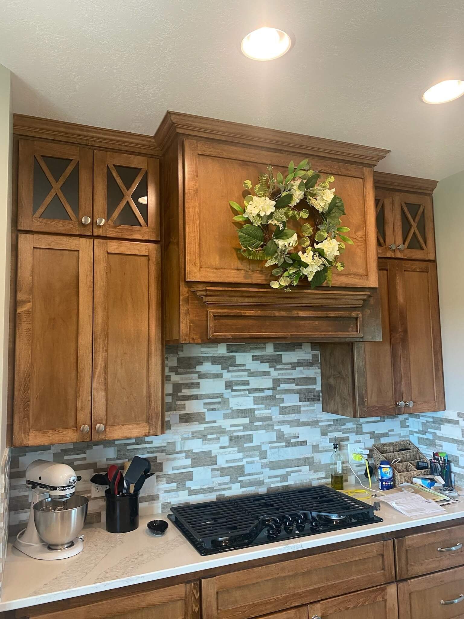 Kitchen stove area with wooden cabinets, a floral wreath on the vent hood, a patterned backsplash, and a stand mixer.