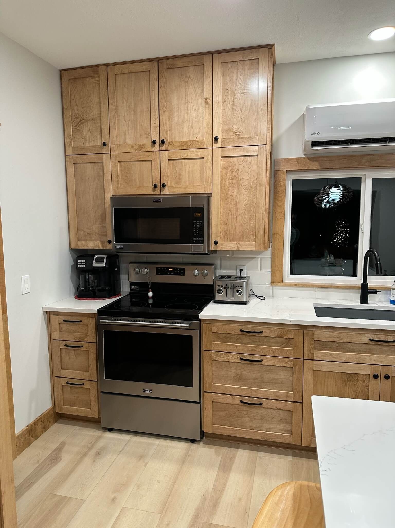 A modern kitchen featuring natural wood cabinets, stainless steel appliances, a white countertop, and a window.