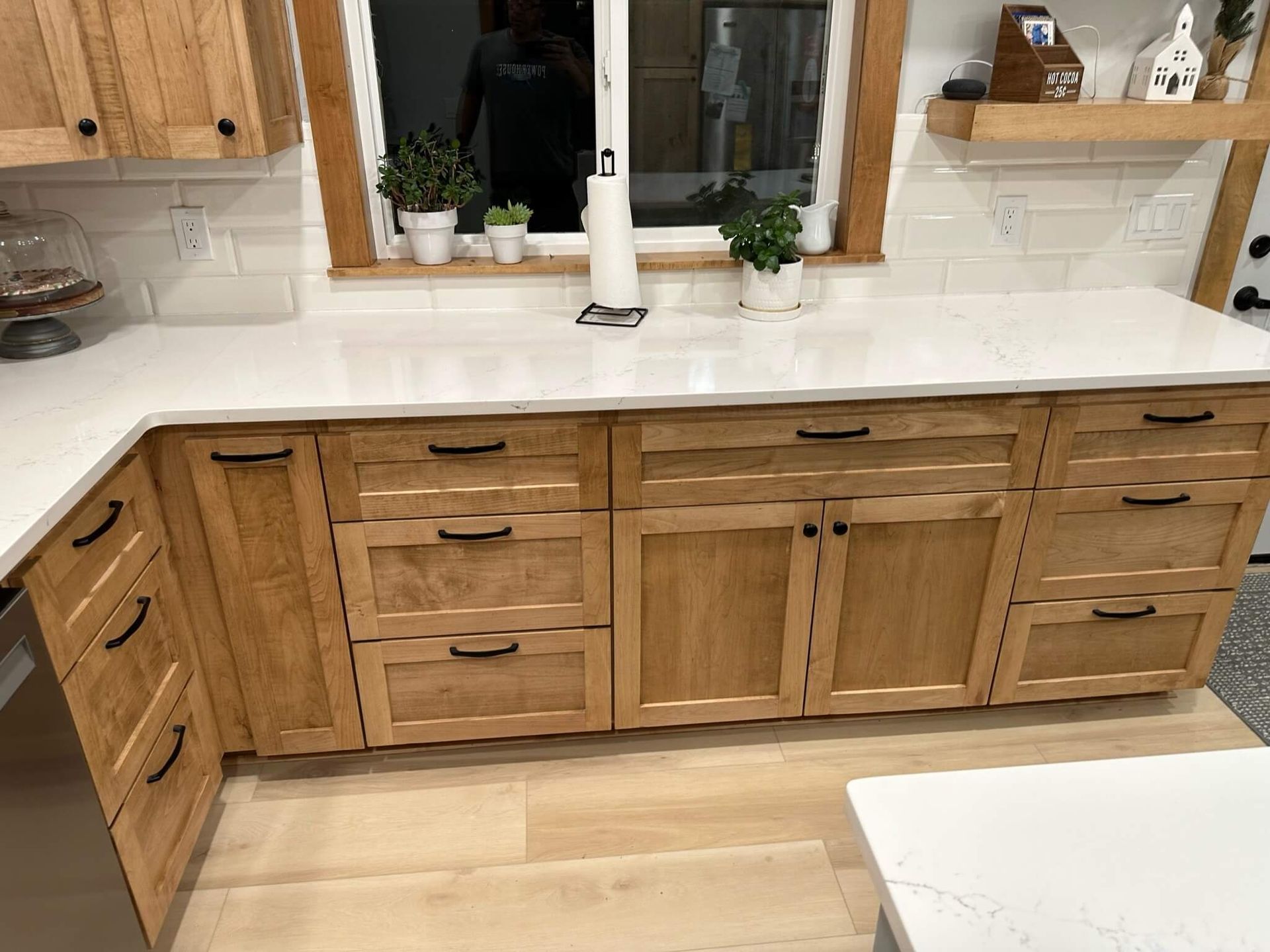 A kitchen counter with light wood cabinets, black hardware, white quartz countertops, and small potted plants by a window.