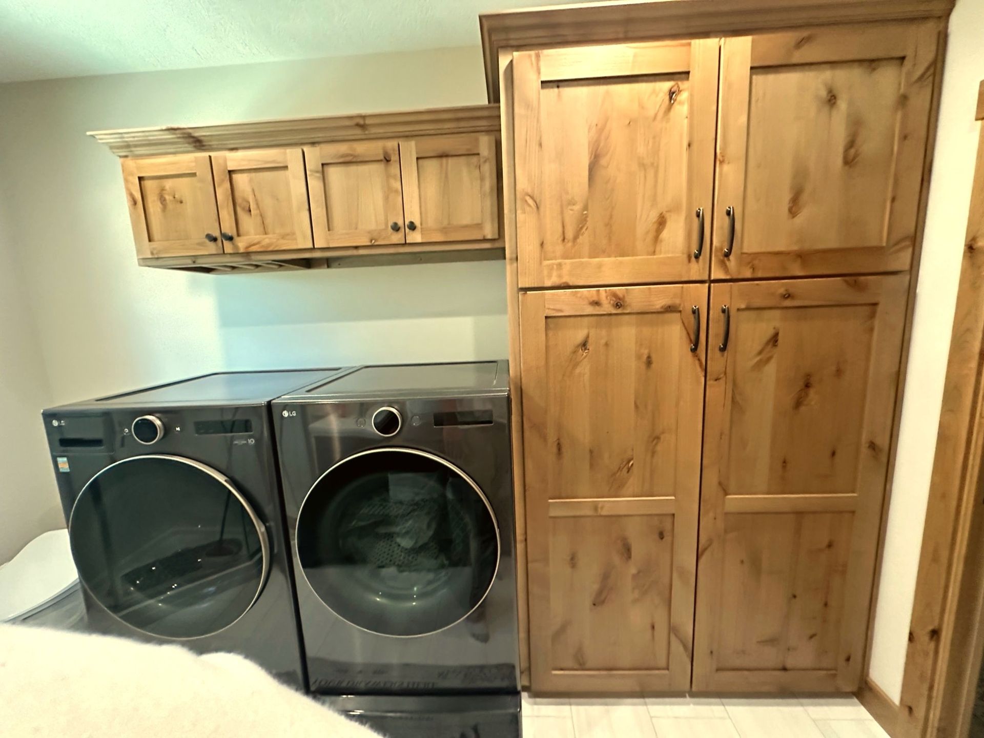 A laundry room featuring two dark grey front-loading machines beneath wooden wall cabinets and next to a tall pantry unit.