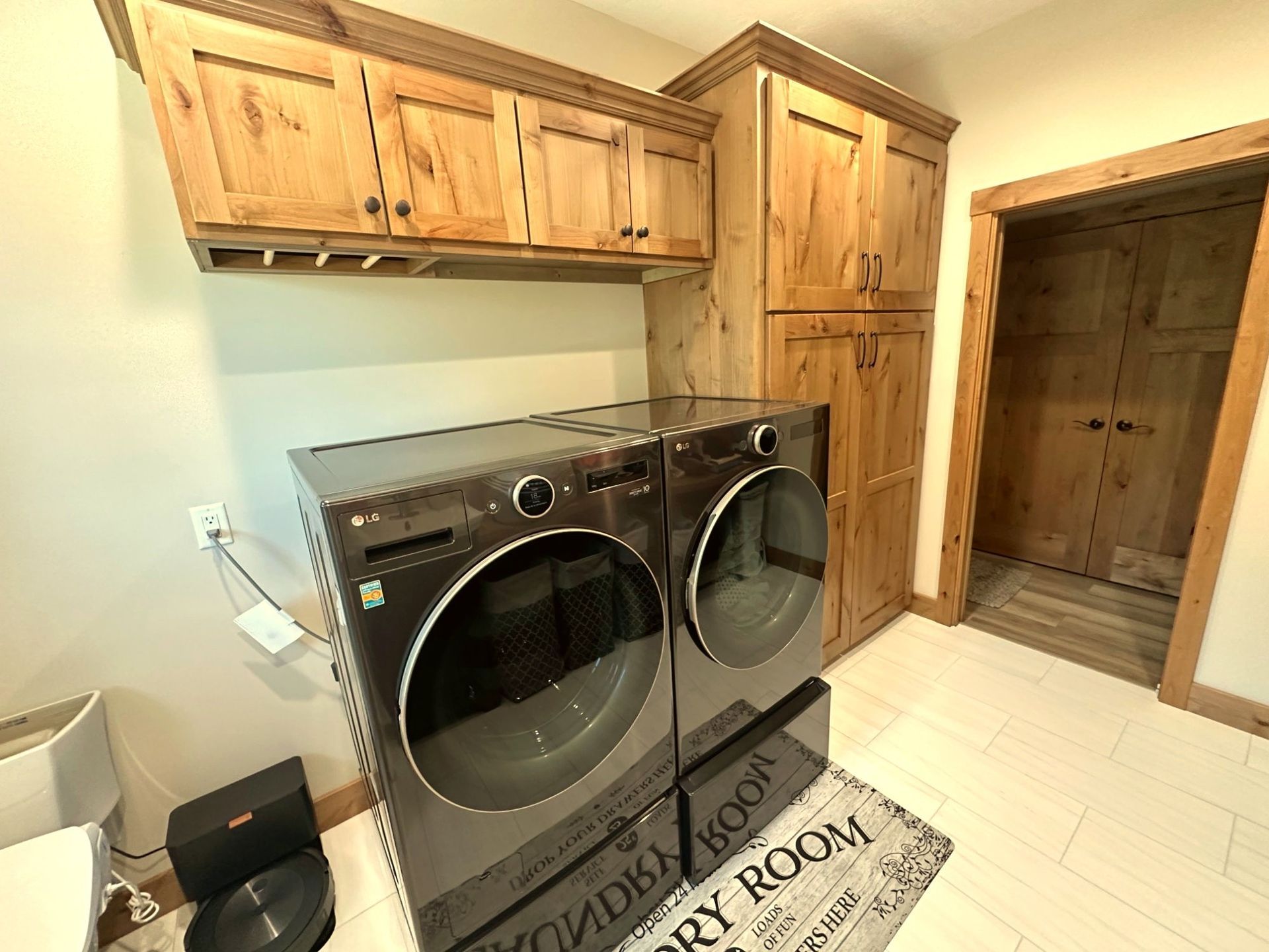 A laundry room with dark grey front-loading machines, wooden cabinets, and a rug on a light tiled floor.