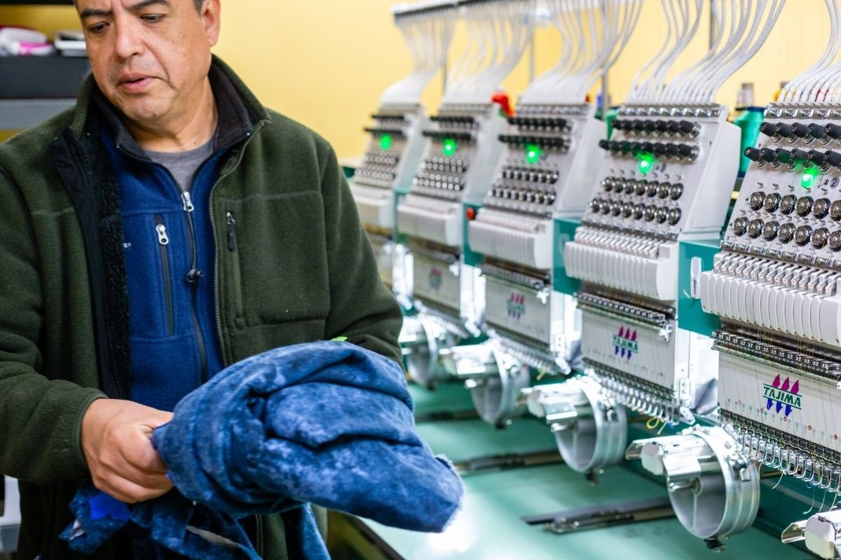 Man holds blue fabric, working with multiple embroidery machines in a workshop.
