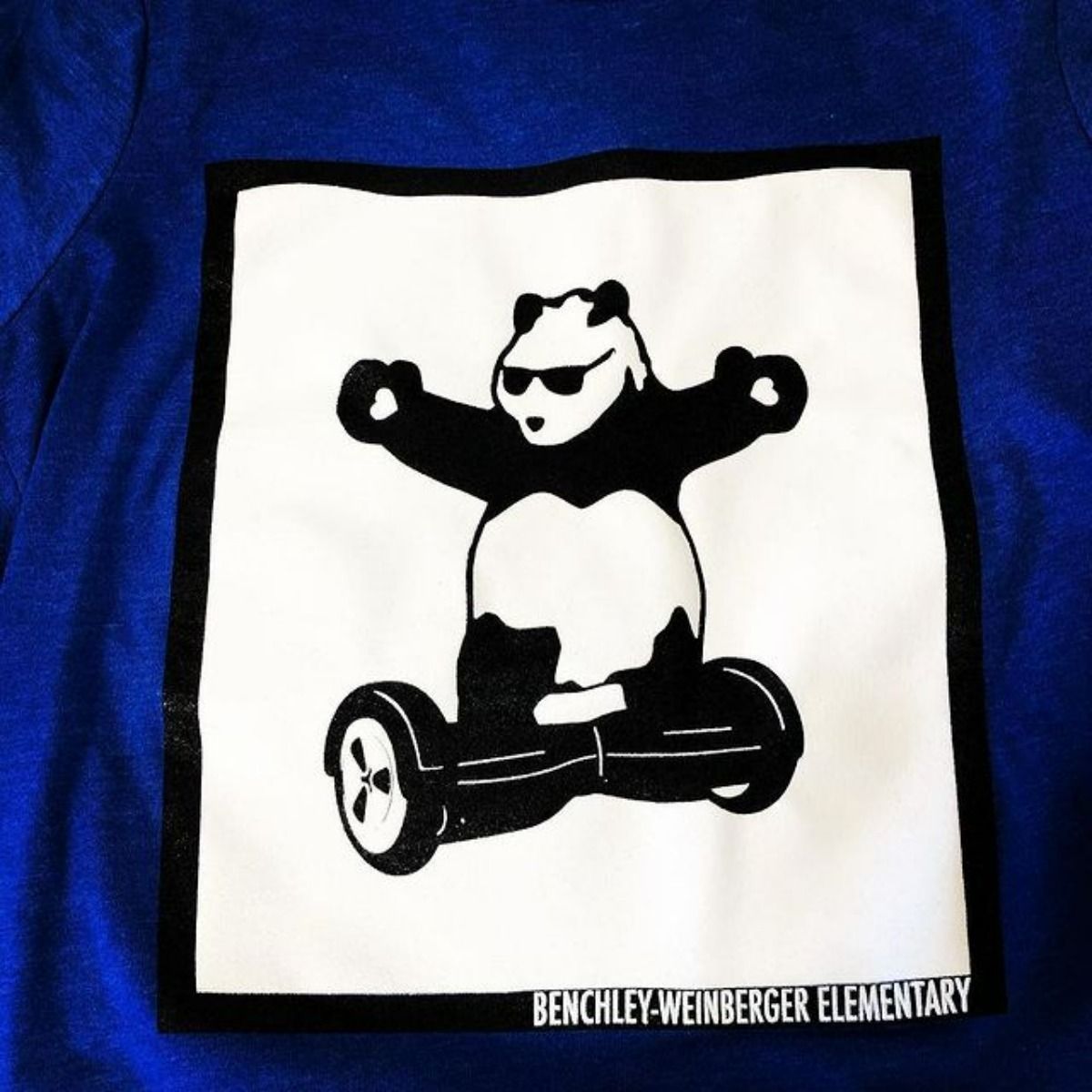 Panda wearing sunglasses, riding a hoverboard. The design is on a blue Benchley-Weinberger Elementary shirt.