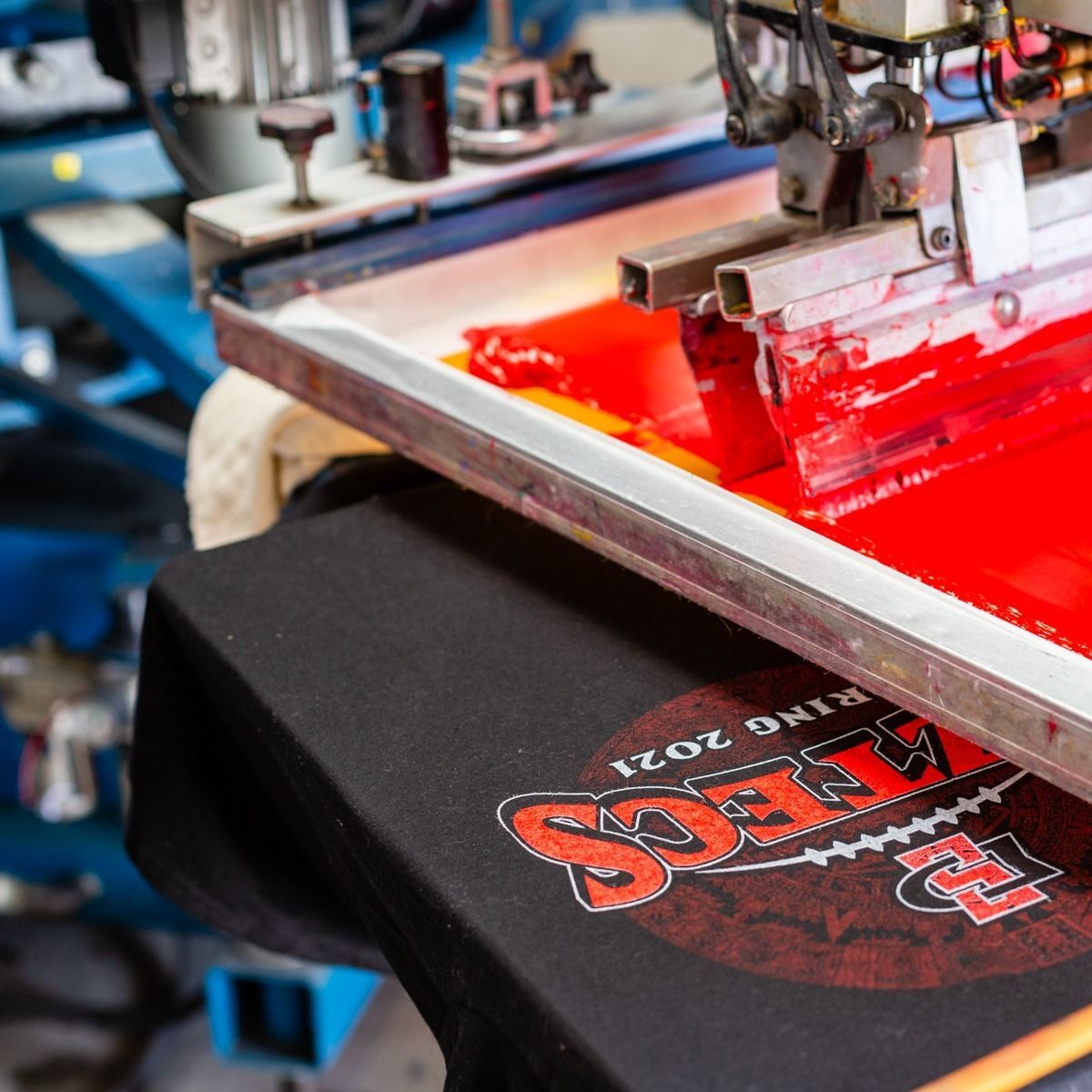 Red ink being screen printed onto a black t-shirt with a design, industrial setting.