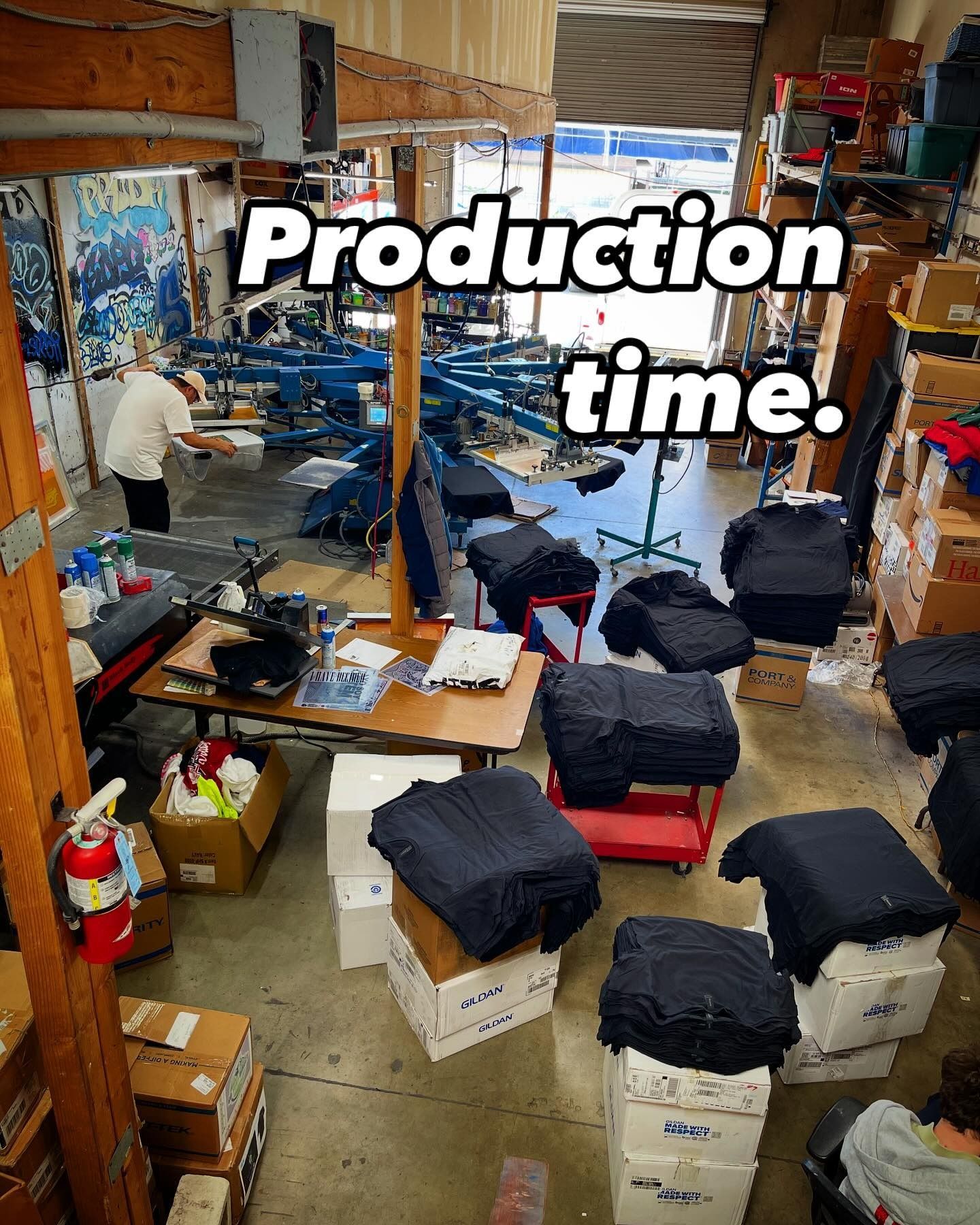 Production area with printing equipment. Black garments on boxes and carts, a person works in the background.