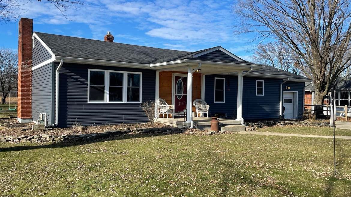 A single-story, dark blue house with a brick chimney and a white-columned front porch, set on a grassy lawn.