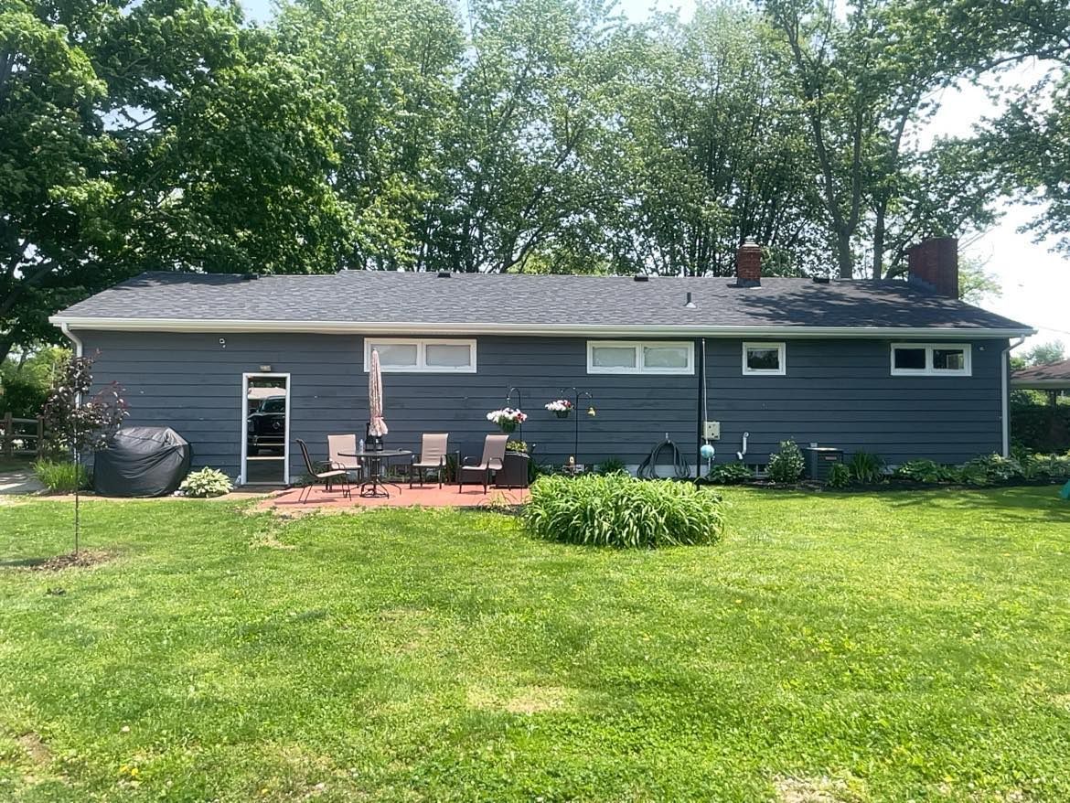 A blue-sided, one-story suburban home with a dark shingle roof, back patio seating, and a large grassy lawn under trees.