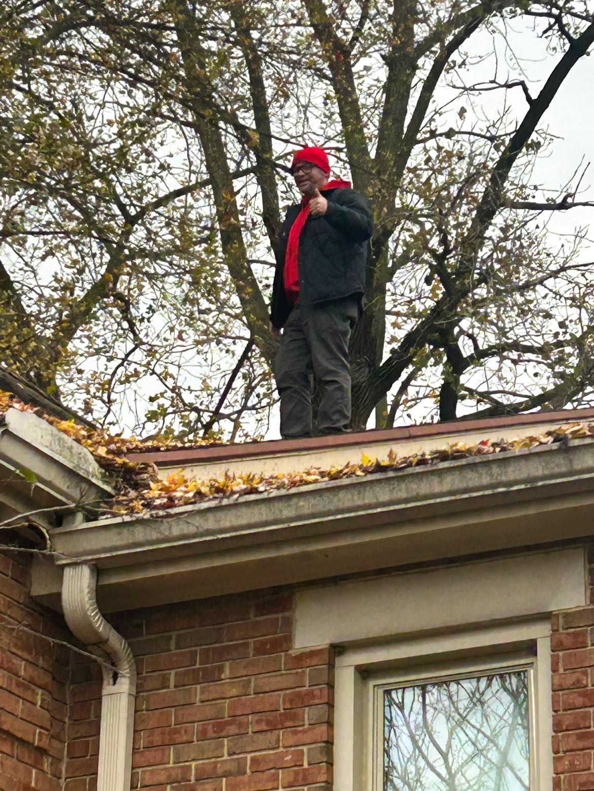 A person in a red hat and jacket stands on a residential roof, giving a thumbs-up against a background of bare trees.
