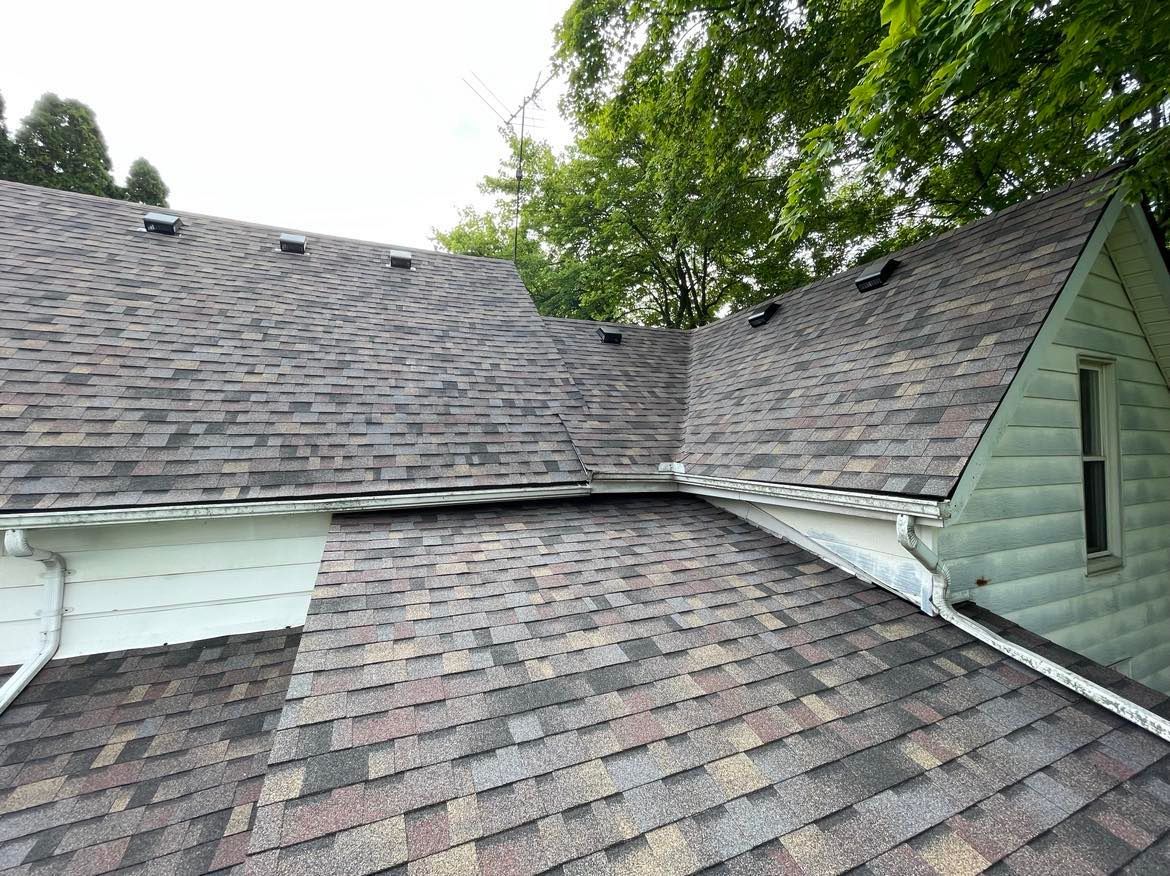 An exterior view of a shingled roof meeting at a valley, with green trees in the background and white siding.