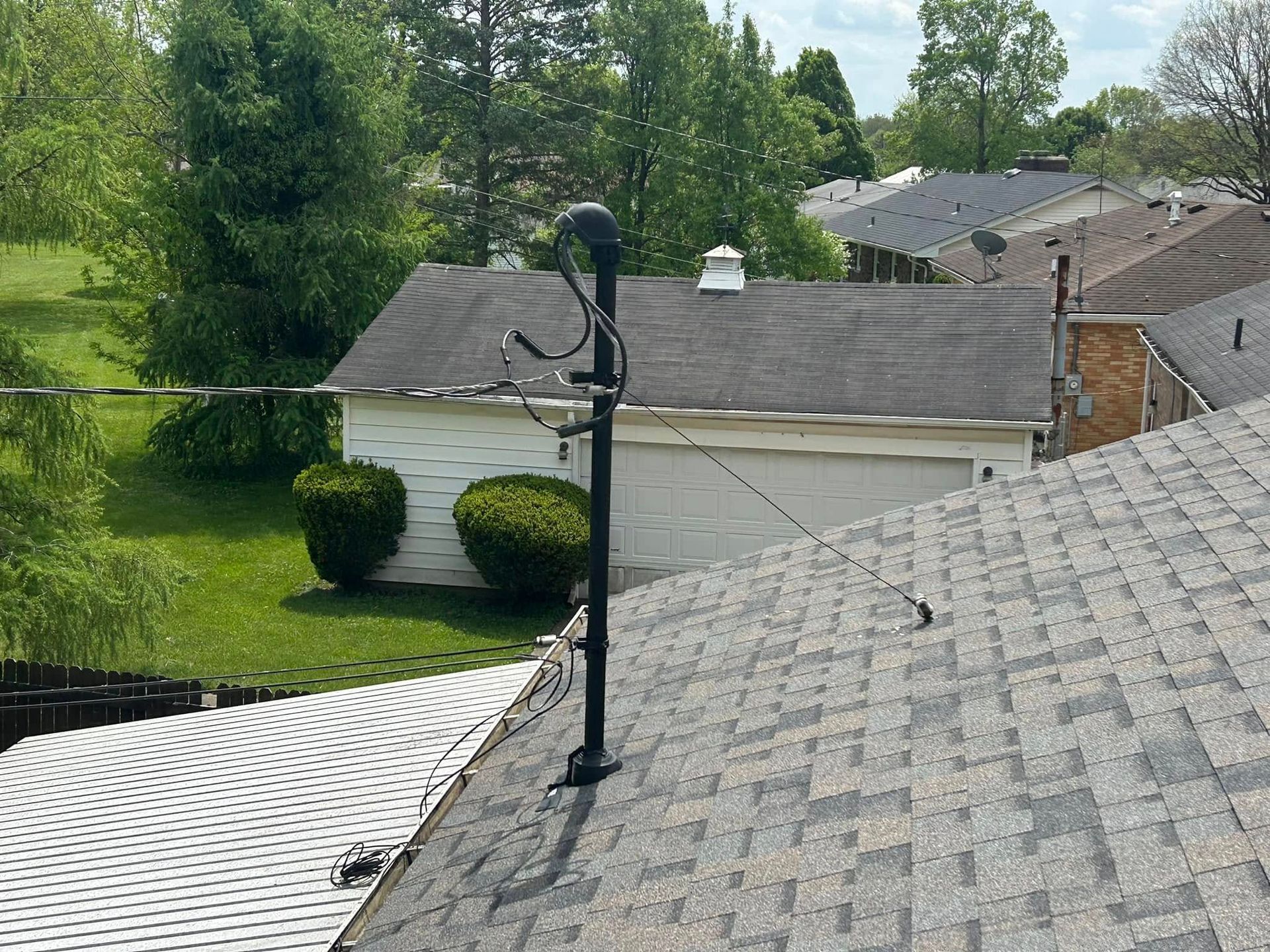 A black mast supporting an electrical service drop wire on a shingled residential roof, with a garage in the background.