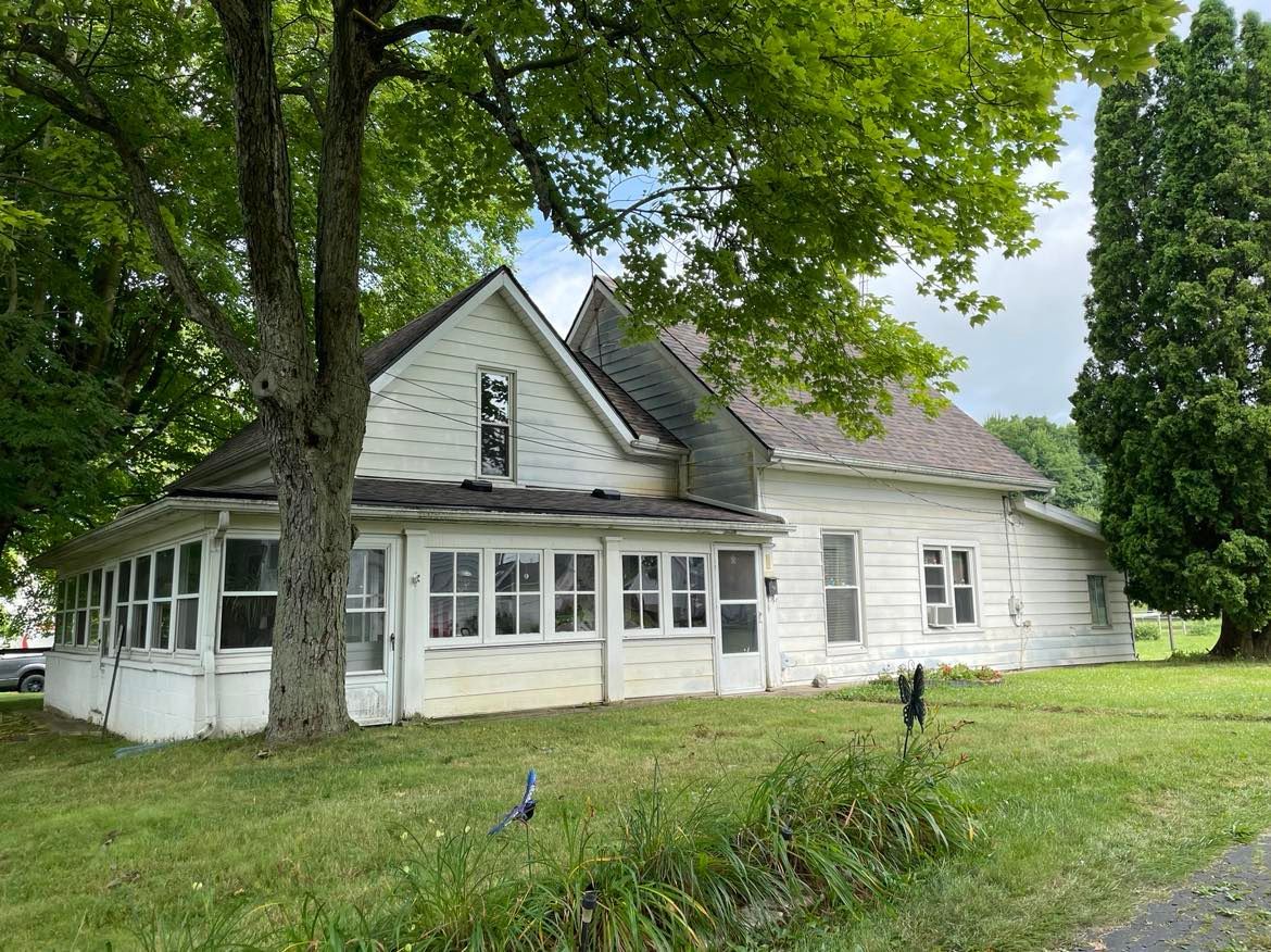 A white, weathered wooden house with a wrap-around enclosed porch, surrounded by green trees and a grassy lawn.