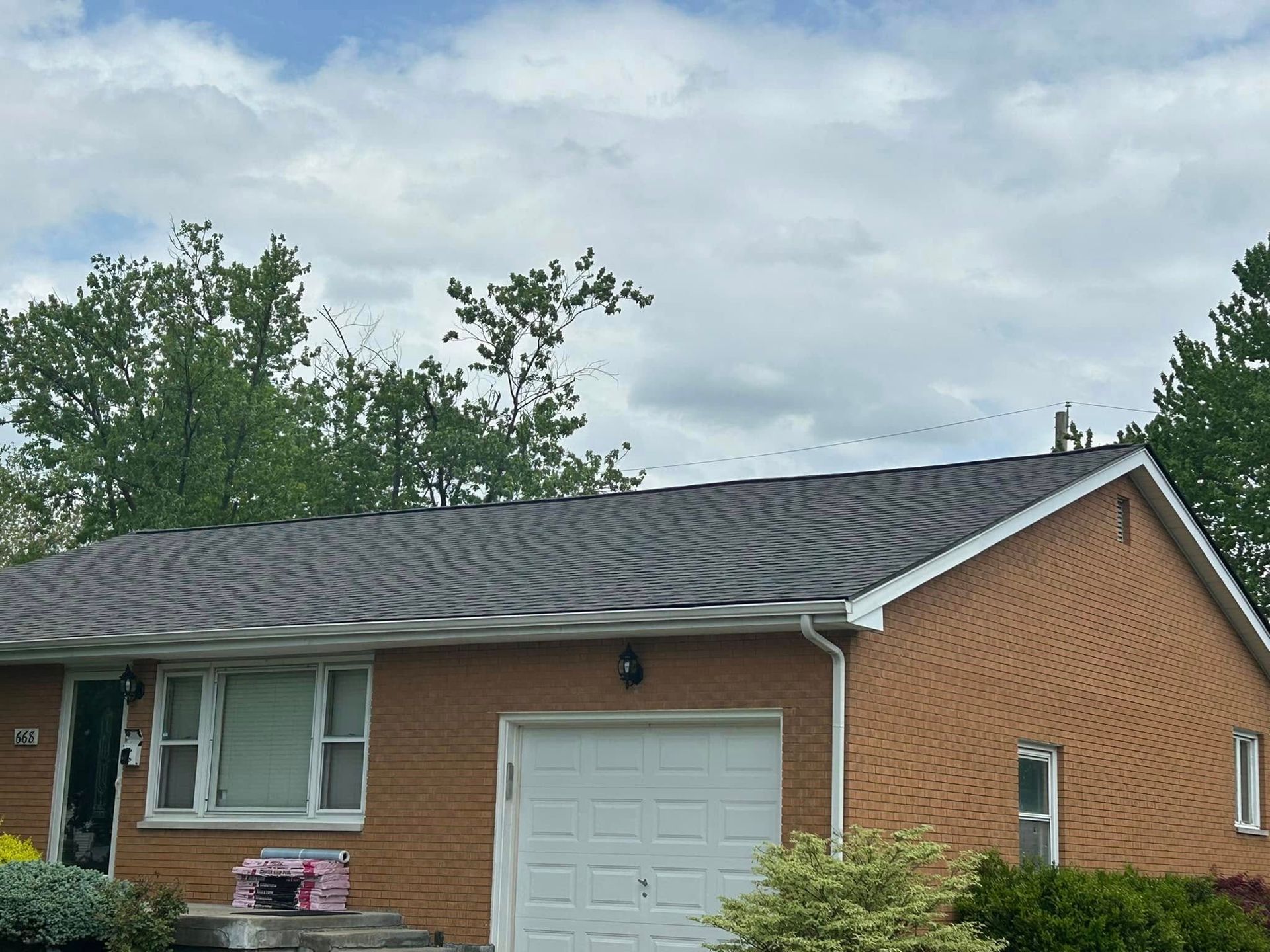 A single-story brick house with a dark gray shingled roof, a white garage door, and a green tree under a cloudy sky.