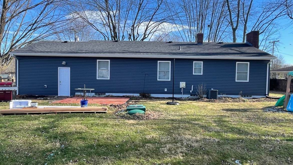 Blue exterior siding of a single-story house featuring several windows, a white door, and a brick chimney in a backyard.