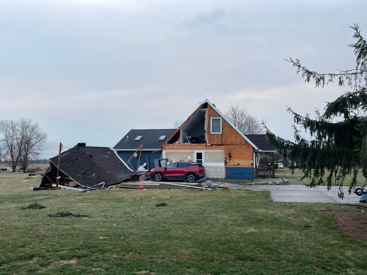 A house with severe storm damage, including a collapsed roof section and exposed interior walls, with a red SUV parked.