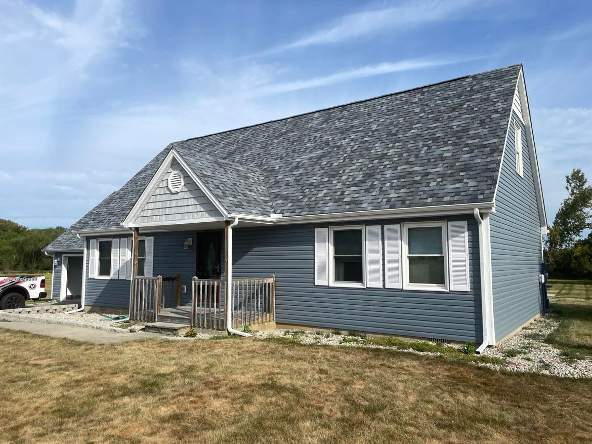 A blue, two-story house with a grey shingled roof, white shutters, and a front porch, set on a dry grass lawn.