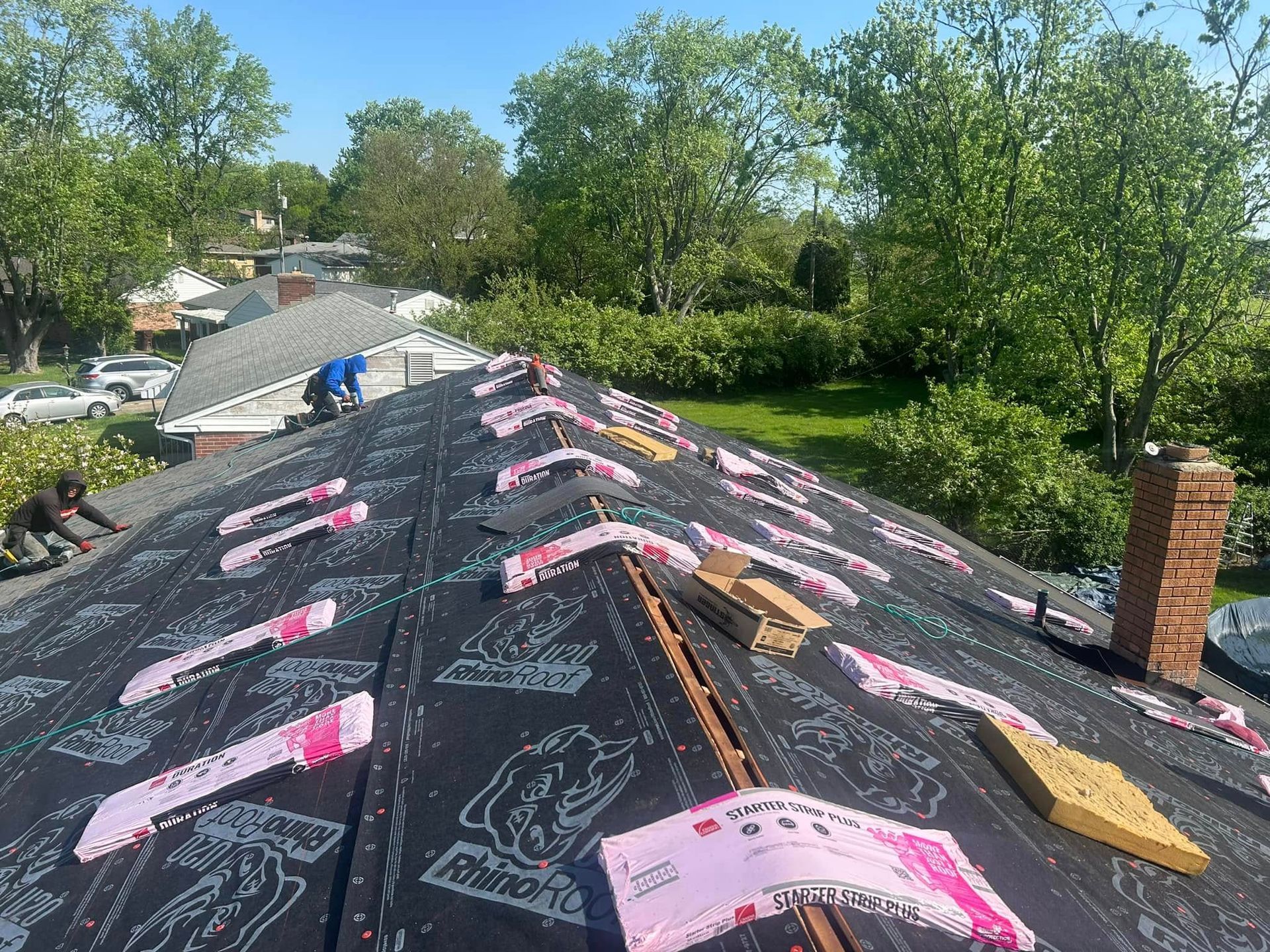 Workers install dark, branded roofing underlayment on a residential roof with a brick chimney in a sunny, green setting.
